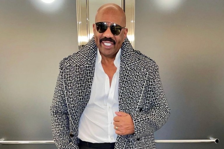 Steve Harvey Wears Balmain Pre-Fall 2021 Monogram Coat Paired With Saint Laurent White Shirt, Black Trousers, and Boots During Latest Style Moment in Paris