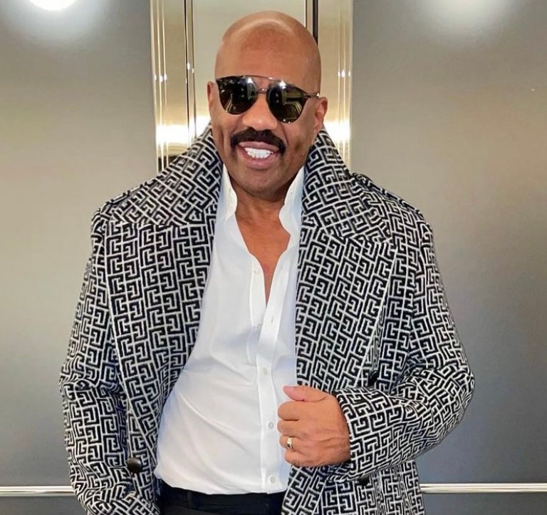 Steve Harvey Wears Balmain Pre-Fall 2021 Monogram Coat Paired With Saint Laurent White Shirt, Black Trousers, and Boots During Latest Style Moment in Paris